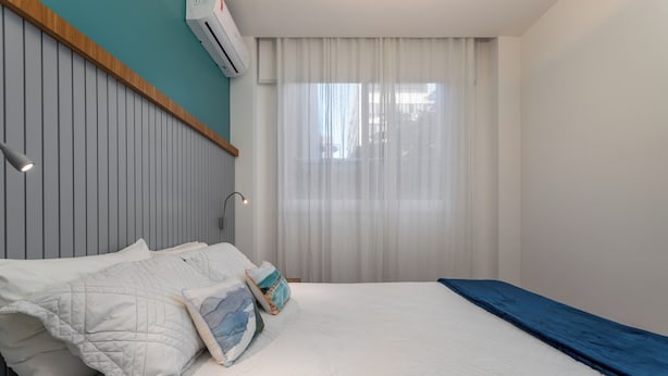 Un lit bien fait, recouvert d’une couvre-lit bleue, avec une tête de lit en bois et une fenêtre ornée de rideaux blancs.