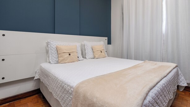 Un dormitorio con una cama, ropa de cama blanca, un plaid color beige y un cabecero blanco.