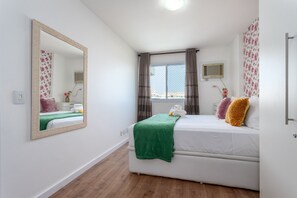 A bedroom with a bed, a mirror, a window with curtains, a wall-mounted air conditioner, and a wooden floor. - Próximo Do Parque Olimpico Bem Localizado | Sa1005 (Rio de Janeiro)