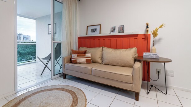 A modern living room with a beige sofa, a red accent wall, a small side table, and a view of the outdoors. - Beleza No Recreio | Para 5 Pessoas | Sa201 (Rio de Janeiro)