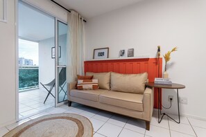 A modern living room with a beige sofa, a red accent wall, a small side table, and a view of the outdoors. - Beleza No Recreio | Para 5 Pessoas | Sa201 (Rio de Janeiro)