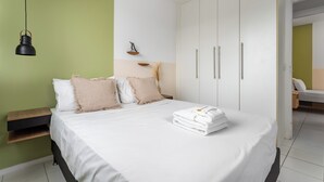 A neatly made bed with white linens and a folded towel, a bedside table with a lamp, and a wardrobe with silver handles. - Beleza No Recreio | Para 5 Pessoas | Sa201 (Rio de Janeiro)