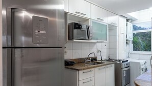 A modern kitchen with stainless steel appliances, white cabinetry, and a view of greenery outside. - Beleza No Recreio | Para 5 Pessoas | Sa201 (Rio de Janeiro)
