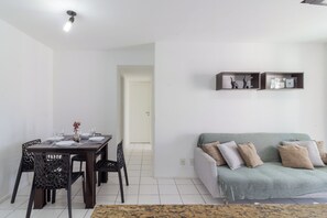 A dining area with a table set for four, a sofa with cushions, and a wall-mounted shelf with decorative items. - Apartamento Com Varanda No Recreio | Sa907 (Rio de Janeiro)