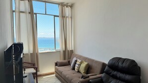 A living room with a sofa, a TV, and a view of the sea through a window. - Refúgio Em Salvador Com Vista Para O Mar (Salvador)