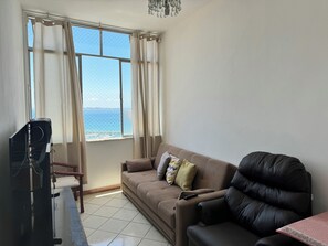 A living room with a sofa, a TV, and a view of the sea through a window.