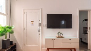 A room with a wooden floor, a white door, a flat-screen TV mounted on the wall, a small white cabinet, and a potted plant. - Sofisticado A Poucos Passos Da Lagoa | Sb301 (Rio de Janeiro)