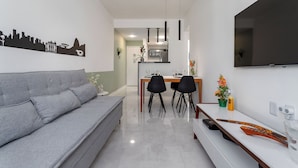 Un soggiorno moderno con un divano grigio, un tavolo da pranzo con sedie, una TV a schermo piatto montata a parete e una cucina sullo sfondo.