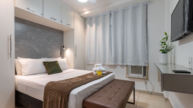 A modern bedroom with a bed, bedside table, bench, and a TV mounted on the wall. - Em Frente Ao Metrô E 8 Minutos Da Praia | Sf228 (Rio de Janeiro)