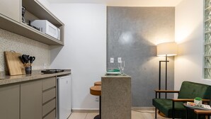 A compact kitchen with a microwave, a small refrigerator, a wooden cutting board, and a set of knives. There is a green upholstered chair and a table with a lamp. - Em Frente Ao Metrô E 8 Minutos Da Praia | Sf228 (Rio de Janeiro)