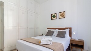 Una cama bien hecha con colcha blanca y marrón, una mesita de noche de madera con lámpara, y dos hojas enmarcadas en la pared.