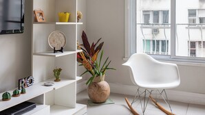 A modern living room with a white sofa, a glass coffee table, a white rocking chair, a white shelving unit with decorative items, and a large window with a view of buildings. - Para Famílias 3Min Da Praia De Copacabana | Sl501 (Rio de Janeiro)
