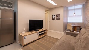 A modern living room with a flat-screen TV on a wooden stand, a sofa, a dining table with chairs, and a kitchen area with a refrigerator. - 3 Minutos Da Praia De Copacabana | Stc303 (Rio de Janeiro)