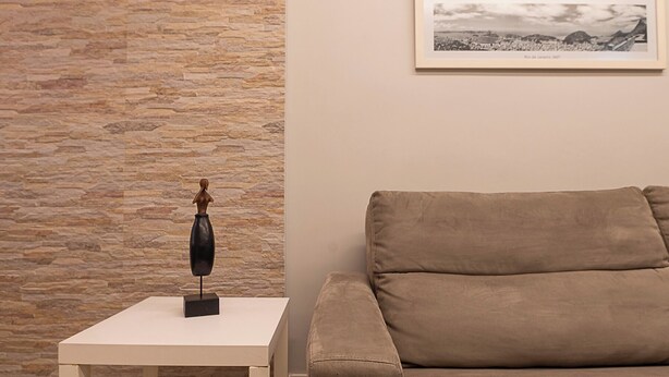 A room with a stone wall, a framed picture, a beige sofa, and a small table with a sculpture. - 3 Minutos Da Praia De Copacabana | Stc303 (Rio de Janeiro)