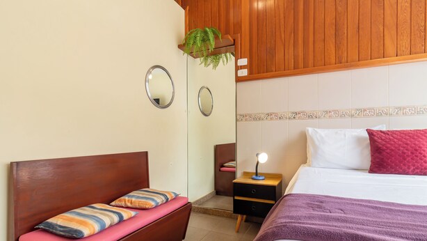 A bedroom with a wooden headboard, a bed with a white and pink pillow, a bedside table with a lamp, and a small plant on the wall. - 5 Minutos Da Praia De Copacabana | Stc905 (Rio de Janeiro)