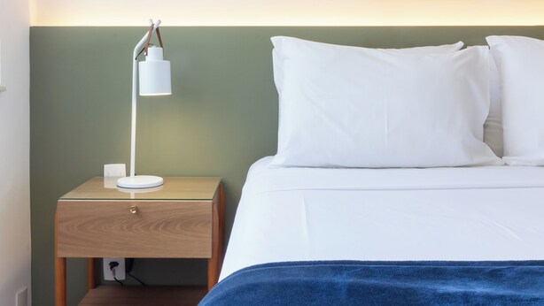 A neatly made bed with white linens and a blue blanket, a bedside table with a white lamp, and two framed pictures on the wall. - Aconchegante A Poucos Minutos Da Lagoa | Vlp412 (Rio de Janeiro)