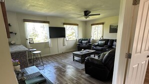 A spacious living room with a wooden floor, a ceiling fan, a black leather sofa, a coffee table, and a TV mounted on the wall. - 3-bedroom House in Ninilchik by Deep Creek (Ninilchik)