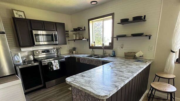 A modern kitchen with dark cabinetry, a central island with stools, a built-in oven, and a window above the sink. - 3-bedroom House in Ninilchik by Deep Creek (Ninilchik)