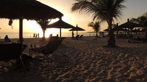 A beach scene with thatched umbrellas, lounge chairs, and palm trees. - Villa pool, residence pools, beach, seaside, beach restaurant (Saly,Région de Thiès)