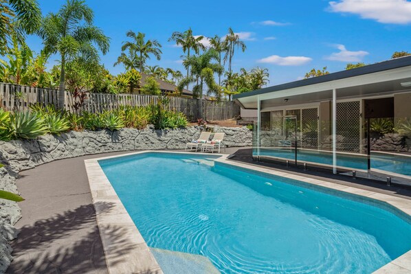 A swimming pool with a wooden deck and lounge chairs, surrounded by tropical plants and a stone wall.