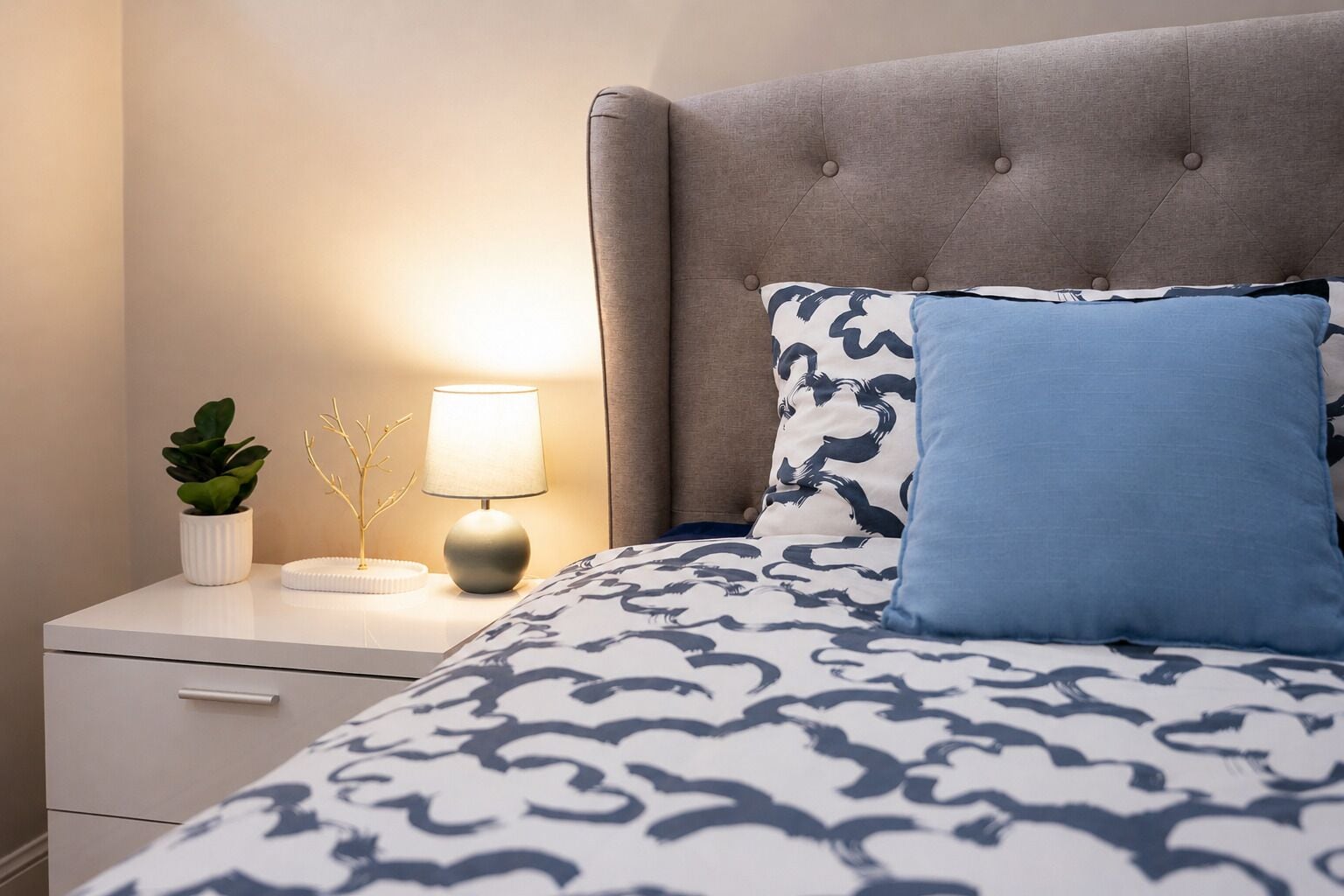 Ein ordentlich bezogenes Bett mit einem gemusterten Überwurf, einem gepolsterten Kopfteil sowie einem Nachttisch mit Lampe, Pflanze und Dekorationsgegenstand.
