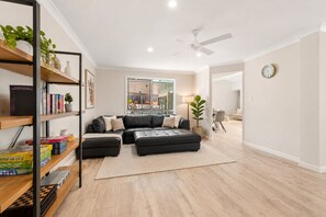 A modern living room with a sectional sofa, a coffee table, a bookshelf, and a large window offering a view of the city. - Modern Family Home – Saltwater Pool, Near Robina (Mudgeeraba)