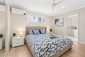 A bedroom with a bed, bedside tables, a ceiling fan, a window with blinds, a painting, and a bathroom door. - Modern Family Home – Saltwater Pool, Near Robina (Mudgeeraba)