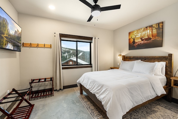 A bedroom with a bed, a ceiling fan, a window with a view of a mountain, and a painting on the wall.