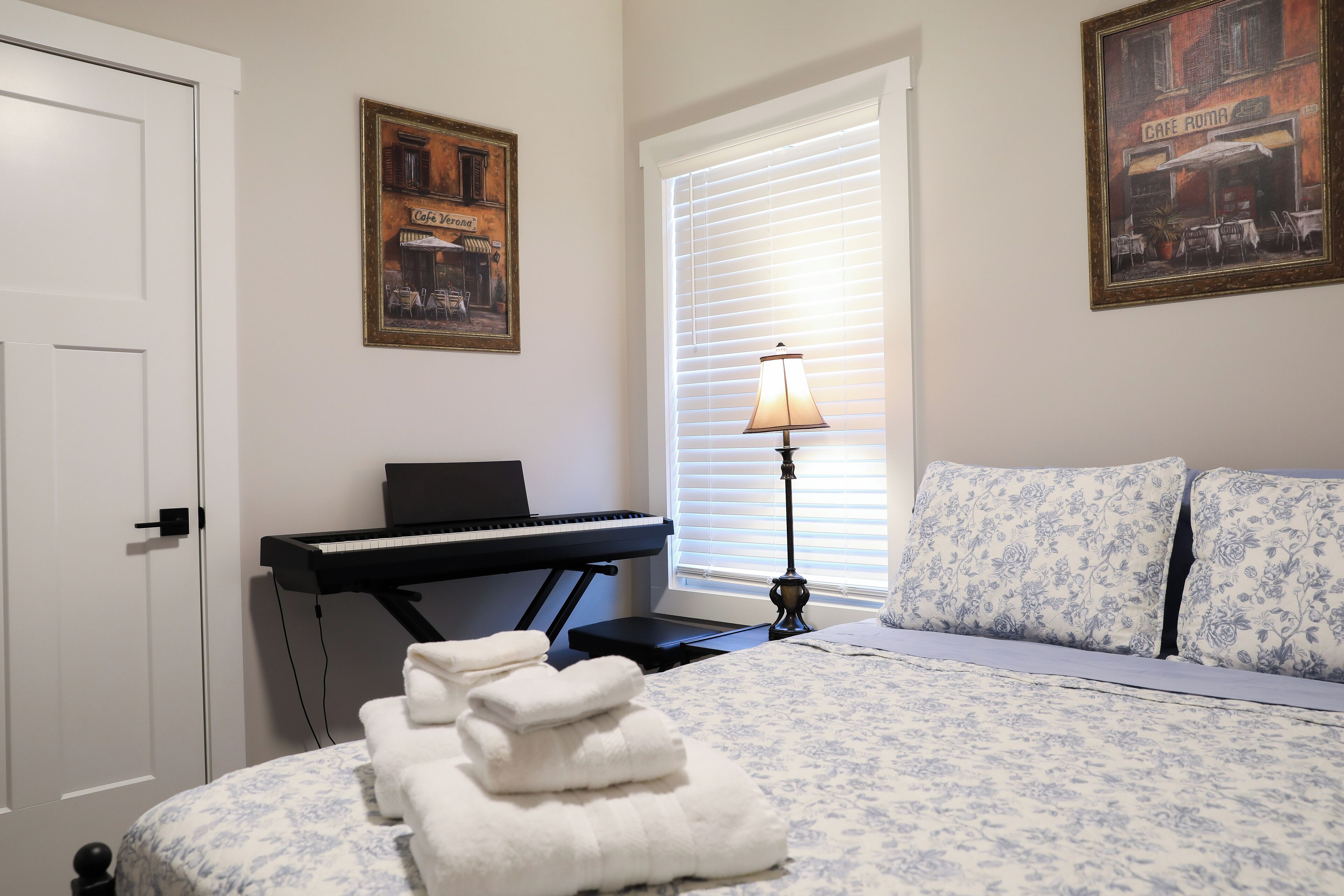 A bedroom with a bed, a piano, a lamp, and two paintings on the wall.