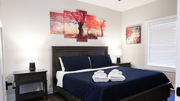 Een keurig opgemaakt bed met een donkerblauwe spreidek en kussens, twee nachtkastjes met lampen, en kunstwerken aan de muur.