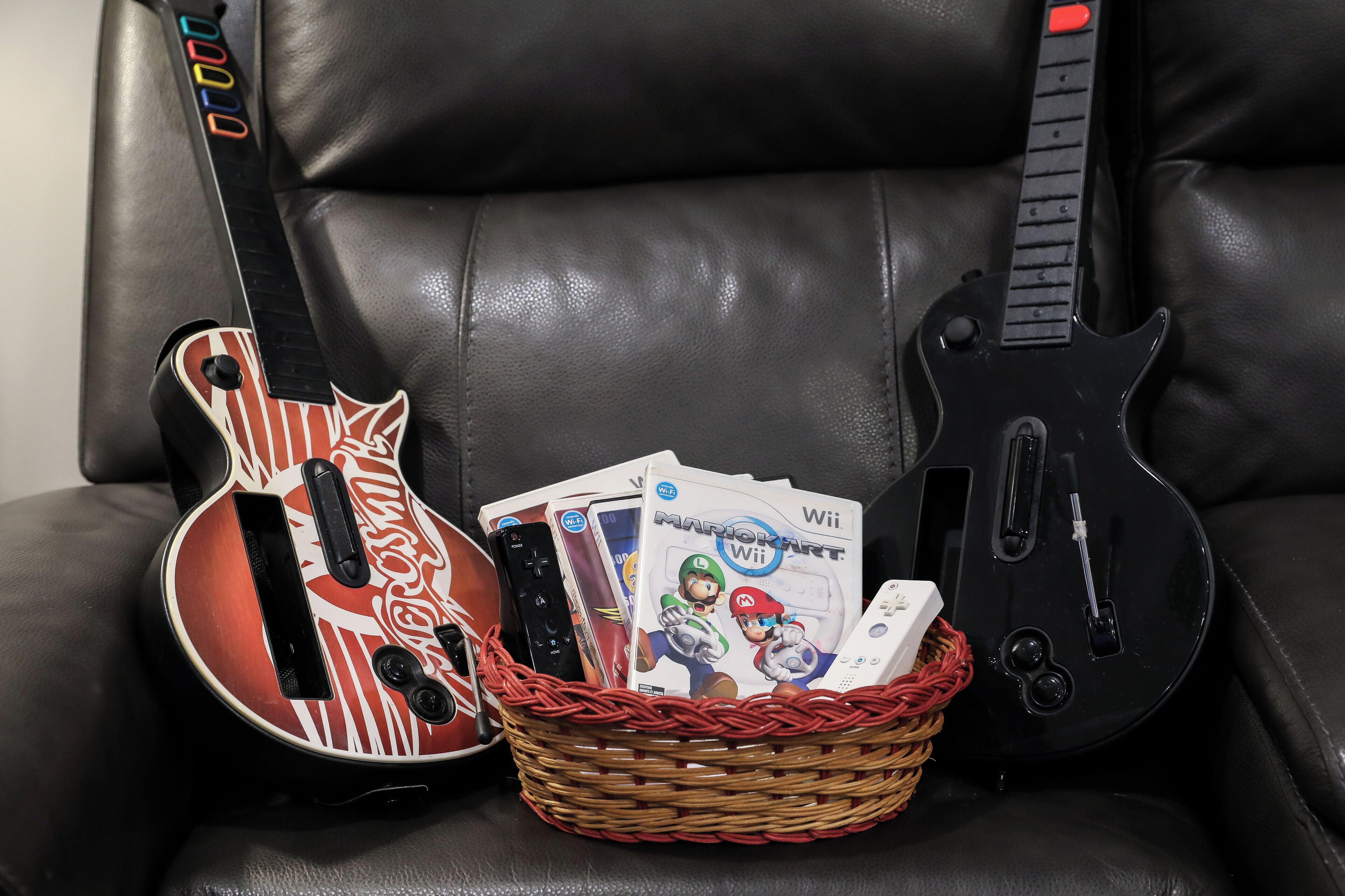 Uma cadeira de couro preto com duas guitarras elétricas e uma cesta com capas de jogos Mario Kart Wii.