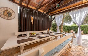 A rustic outdoor dining area with a wooden table set for two, a clock on the wall, and a view of a hedge and trees.