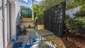 Un pati amb una taula rodona i cadires blaves, una petita zona de jardí amb plantes i un arbre, i una porta mosquitera negra.