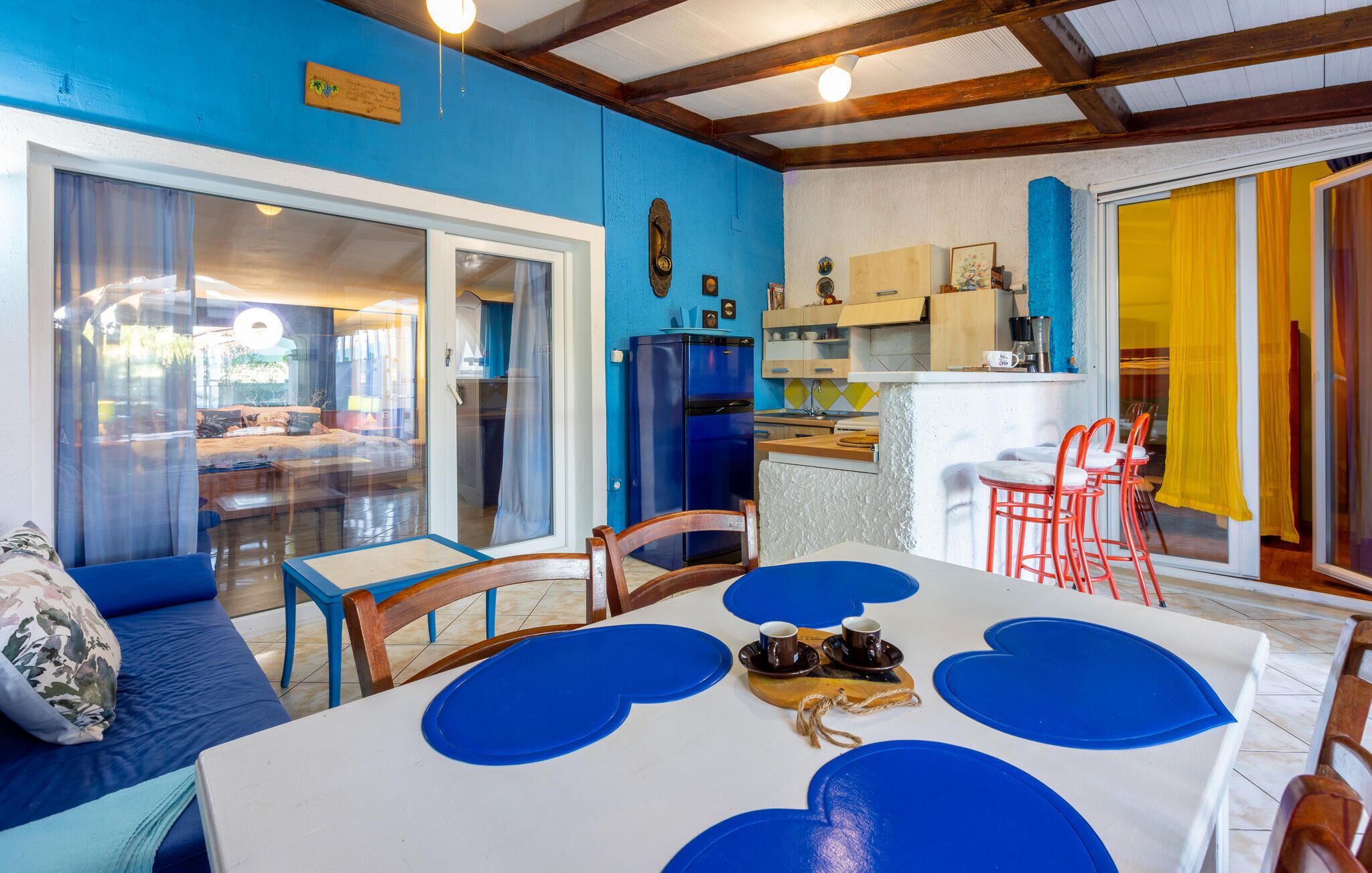 Een eethoek met een tafel gedekt voor vier personen, een blauwe koelkast, en een keuken met houten kastjes en een geel gordijn.