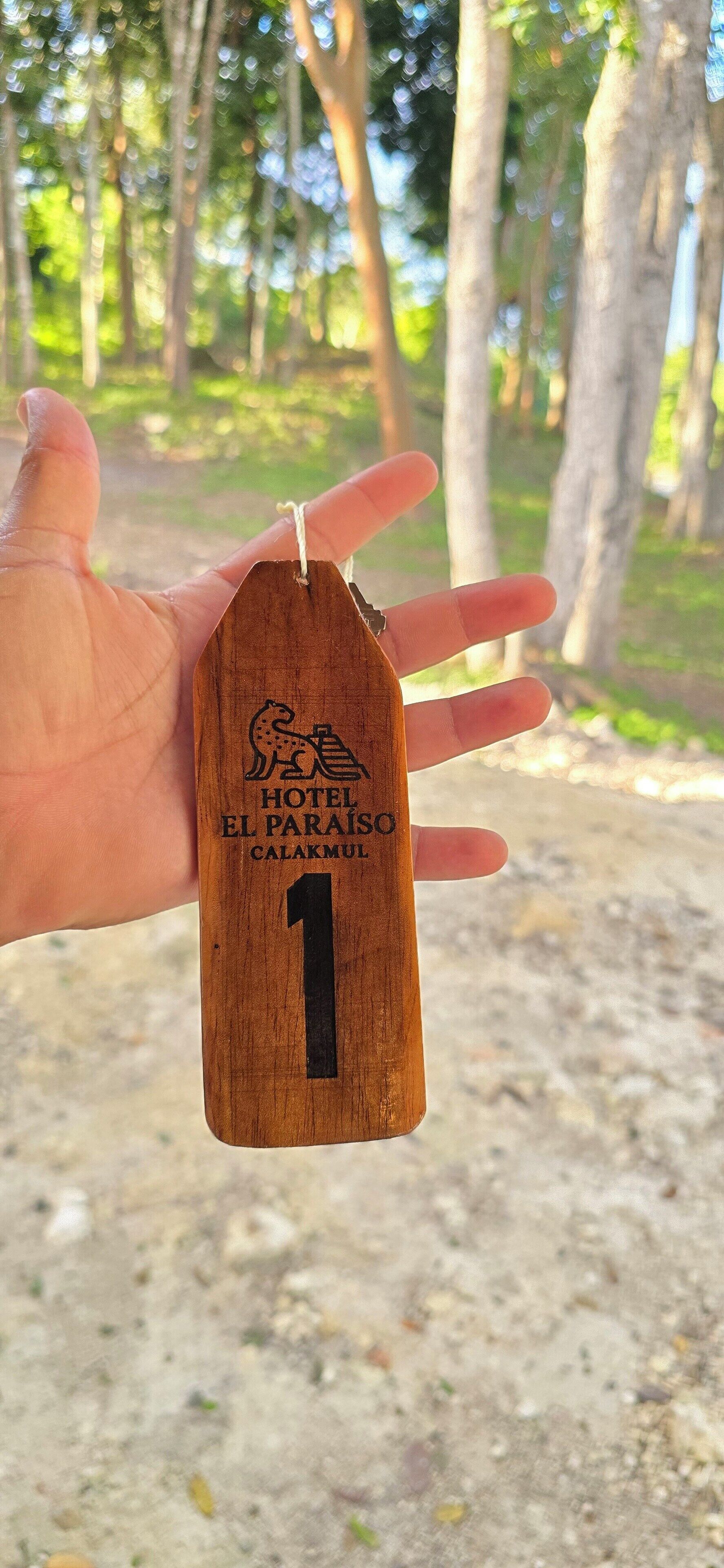 Uma mão segurando uma etiqueta de madeira com o texto 'HOTEL EL PARAISO CALAKMUL 1' e um desenho de leopardo.