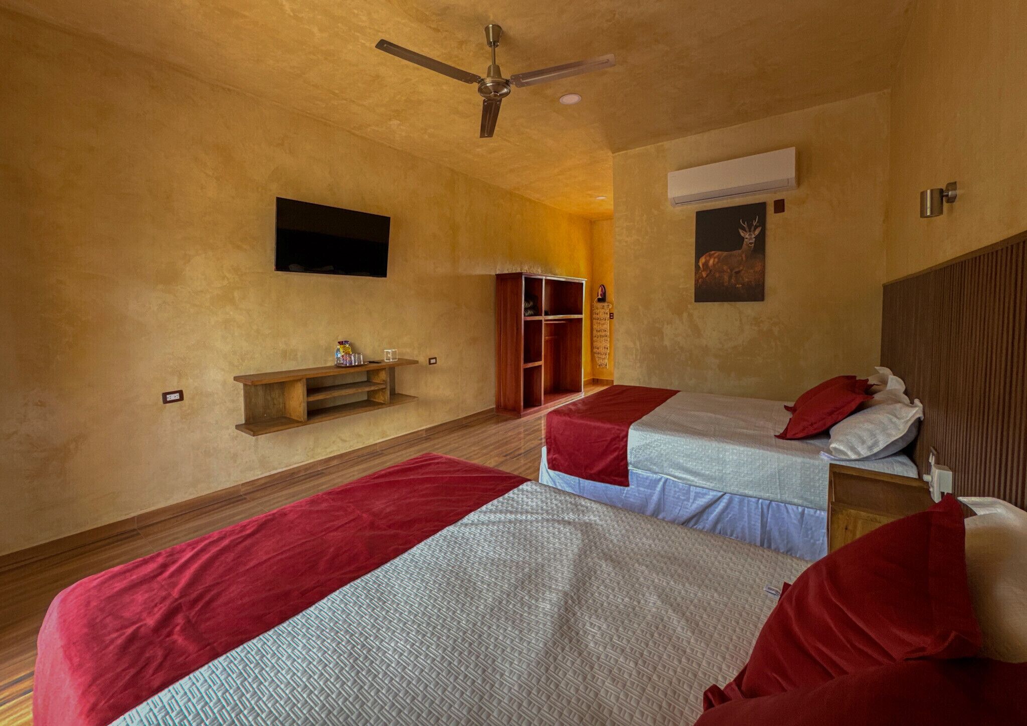 A room with two beds, a wooden bench, a ceiling fan, a mounted TV, and a wall-mounted air conditioner.