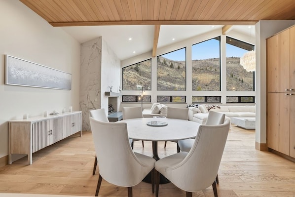 A modern dining area with a round table, white chairs, and a view of a mountainous landscape through large windows.