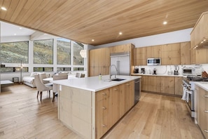 A modern kitchen with wooden cabinets, a large island, and a dining area with a view of the outdoors.