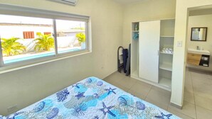 A bedroom with a bed, a wardrobe, a bathroom, and a window with a view of palm trees. - Ocean Breeze Apt #2 - 1BR 2Beds with pool (Oranjestad)