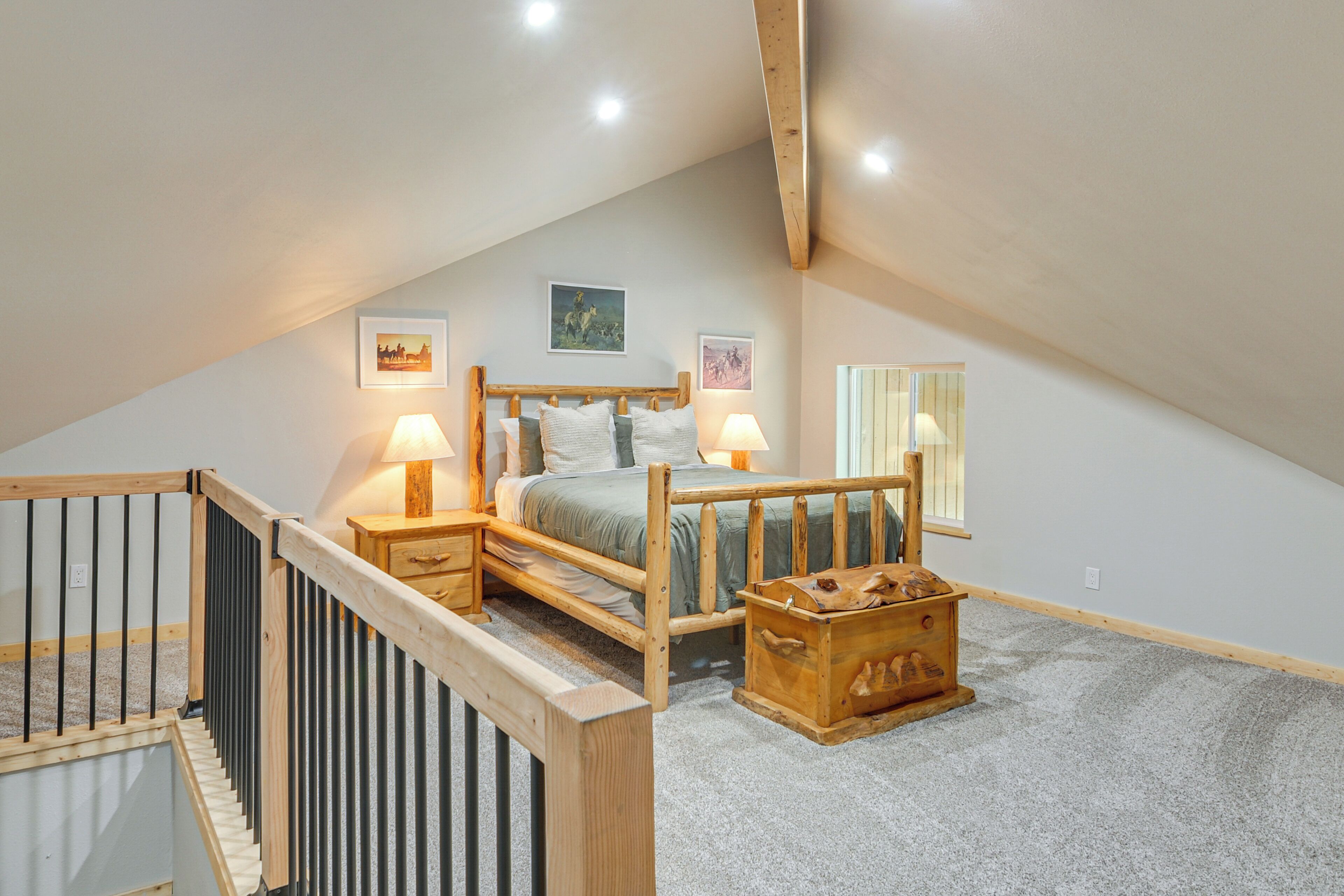 A bedroom with a sloped ceiling, a wooden bed frame, two bedside lamps, a nightstand, a wooden chest, and two framed pictures on the wall.