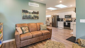 A living room with a brown sofa, a painting on the wall, and a kitchen in the background. - Walk to Historic Chestertown: Dog-Friendly Retreat (Chestertown)