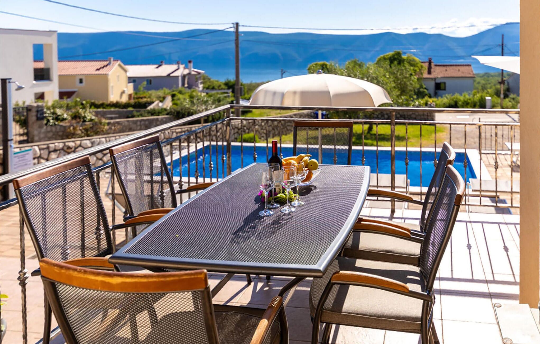 Une table près de la piscine, avec des chaises, une bouteille de vin et des fruits.