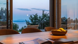 Uma área de refeições com vista para o mar e montanhas, uma tigela de frutas e um livro aberto sobre a mesa.