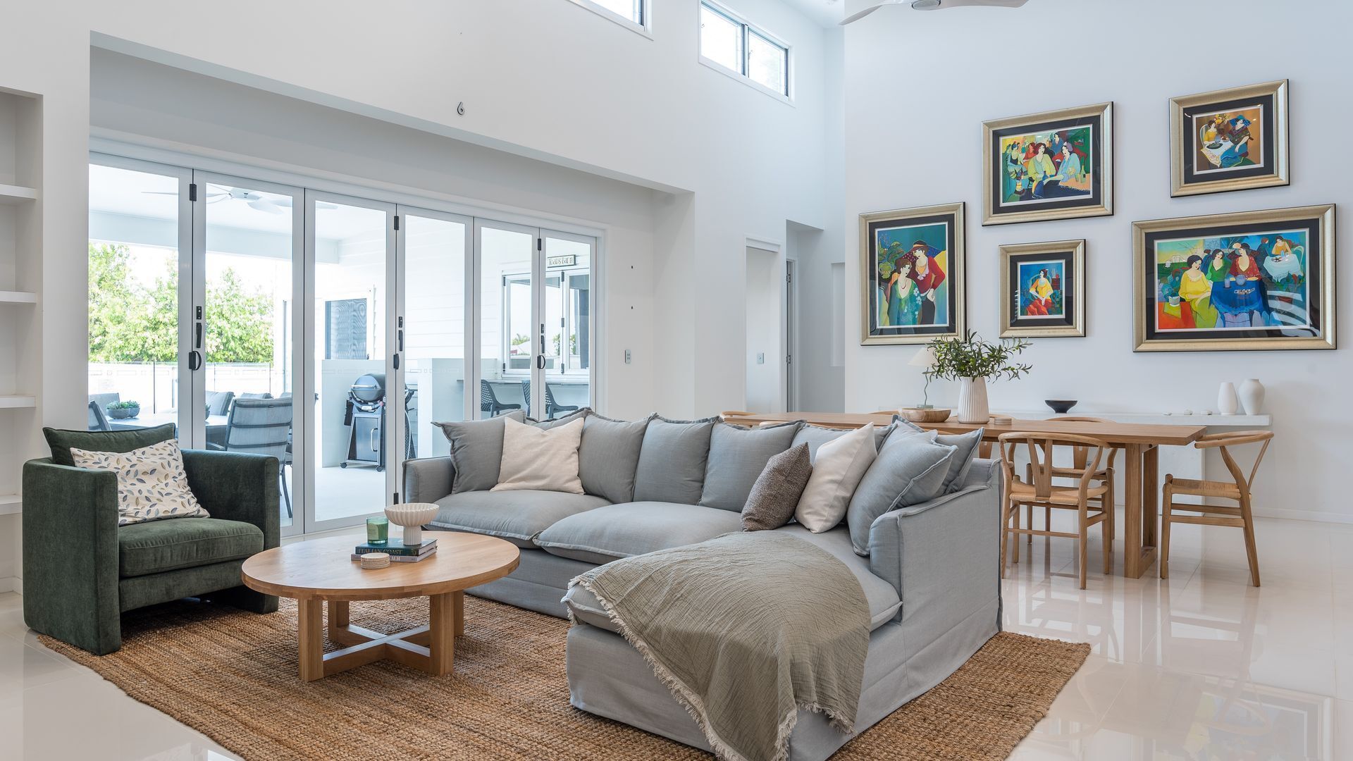 A spacious living room with a grey sofa, a round wooden coffee table, and a dining area with a wooden table and chairs. There are large windows providing natural light and a view of the outdoors.