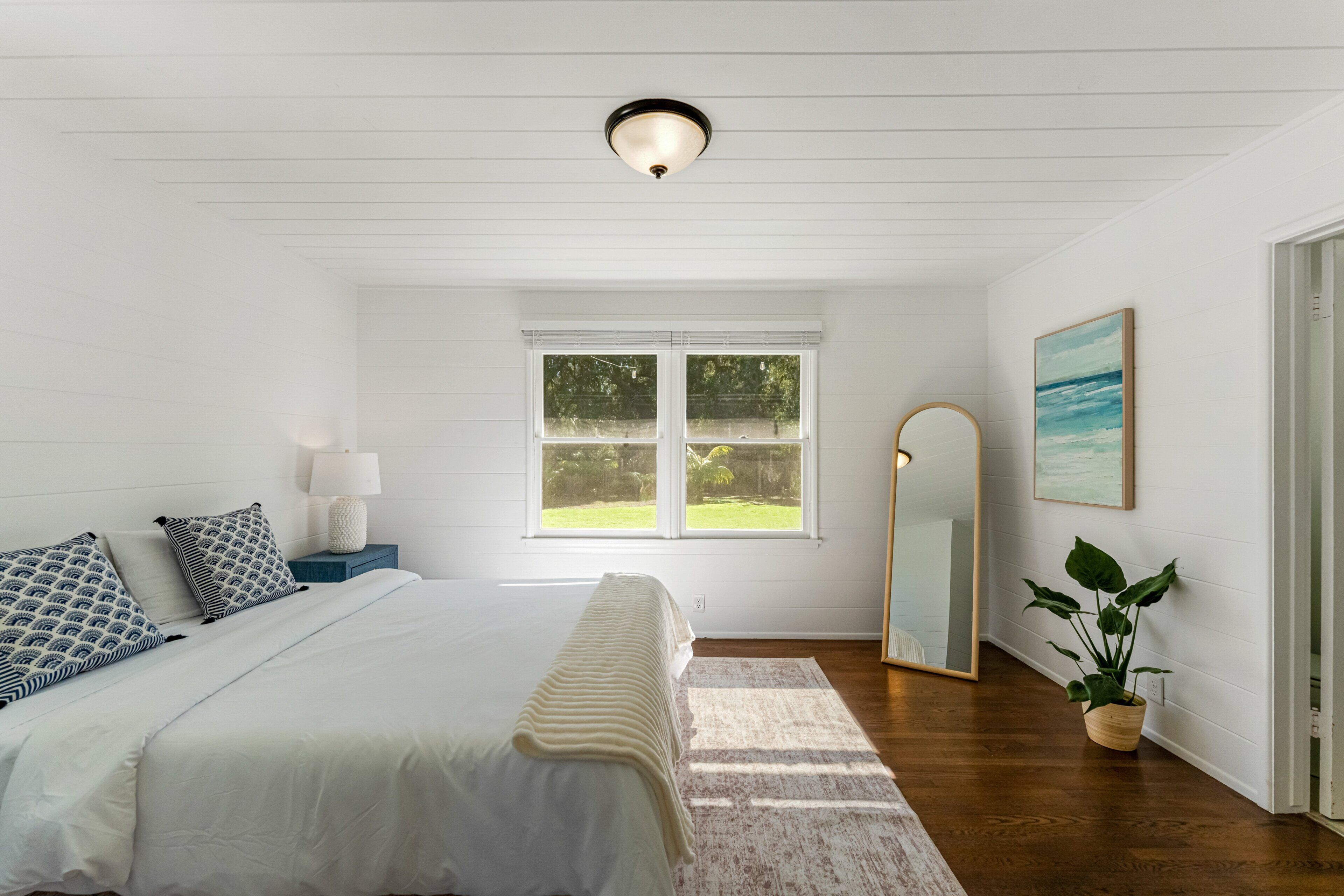 Une chambre à coucher avec un lit, une table de chevet, une lampe, une fenêtre, un miroir et une plante en pot.