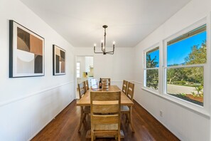 Un espace repas comprenant une table et des chaises en bois, un lustre et de grandes fenêtres donnant sur un paysage verdoyant.