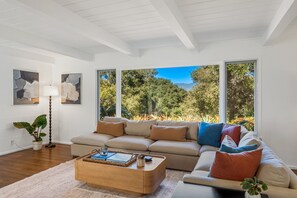 A modern living room with a beige sectional sofa, a wooden coffee table, and large windows offering a view of trees and mountains. - Coastal Farmstead Retreat | The Chosen Collection (Santa Barbara)