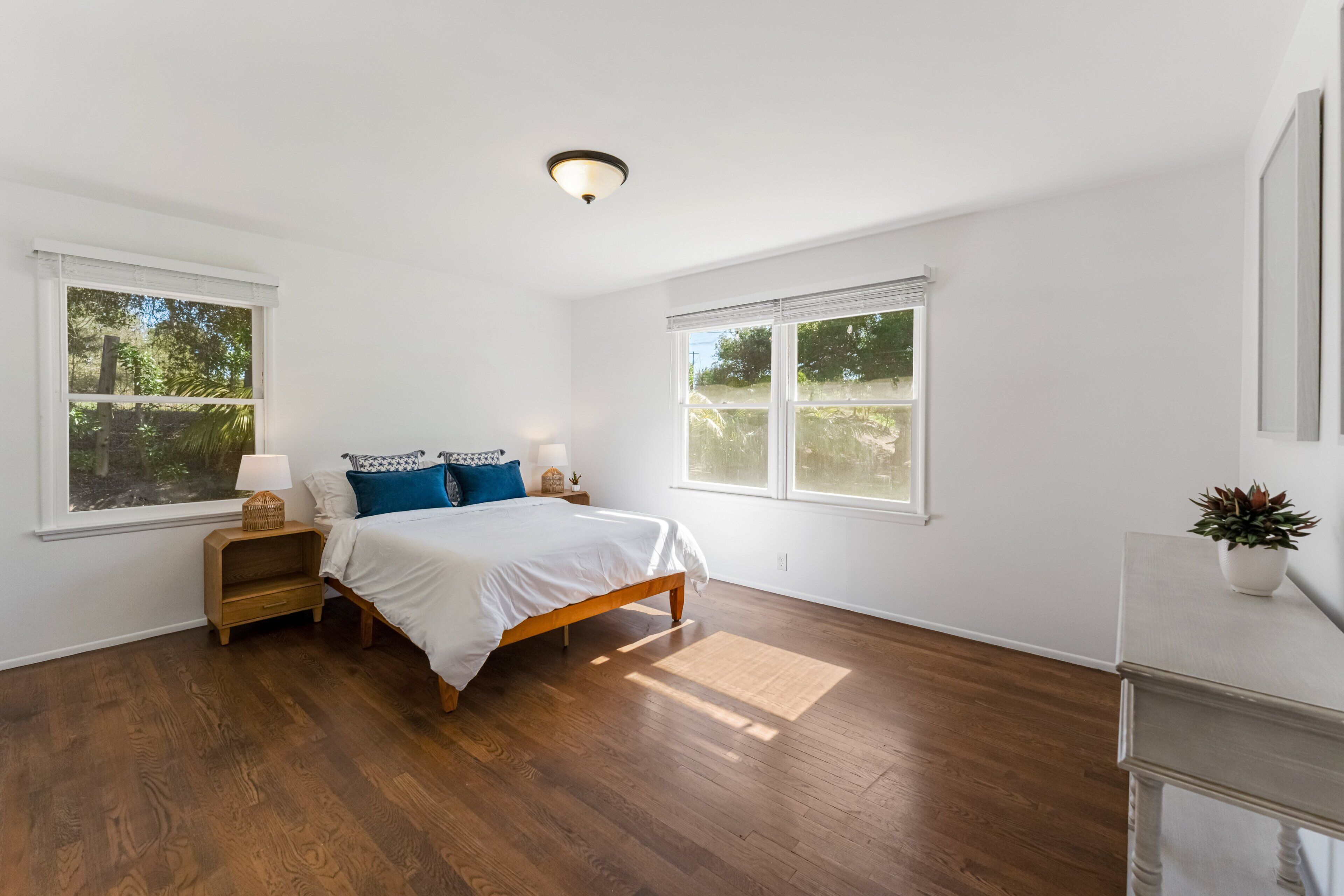 Un dormitorio con una cama de madera, dos ventanas, una mesita de noche, una lámpara y una planta en maceta.