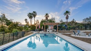 Una piscina rectangular con una pérgola de madera y mobiliario exterior, rodeada de palmeras y una valla.