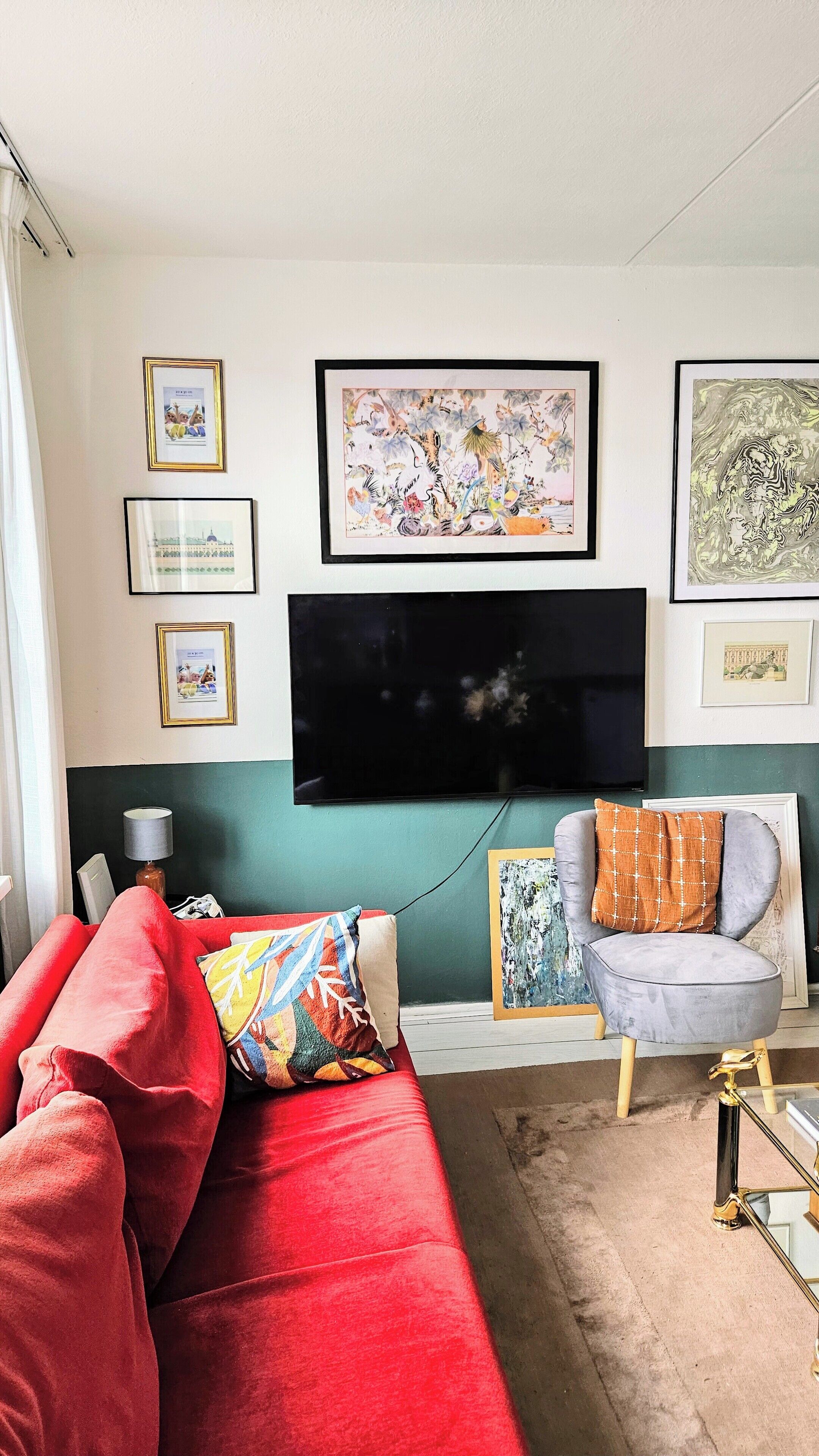 Uma sala de estar com um sofá vermelho, uma poltrona cinza e uma TV fixada na parede. Há quadros nas paredes e um abajur sobre a mesa lateral.